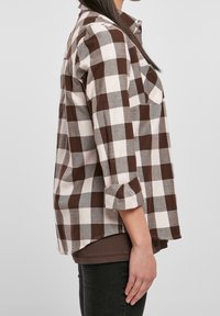 Camicia a quadri marrone e crema con maniche arrotolate, colletto button-down e taschino sul petto. Realizzata in tessuto leggero, presenta un orlo curvo.