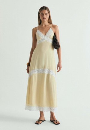 Mujer con un vestido largo de color amarillo pálido con ribete de encaje blanco, bolso negro con cuentas en el brazo, pulsera de madera y sandalias negras.