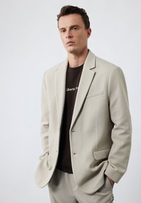 Beżowa marynarka o strukturalnym designie, z dwoma przednimi kieszeniami i zapięciem na jeden guzik, zestawiona z ciemnobrązowym T-shirtem z białym napisem.