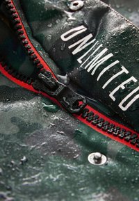 Camouflage zippered fabric with water droplets, featuring a black zipper and red accents. Text reads "UNLIMITED." Texture is smooth.