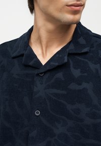 Κοντινό πλάνο ενός ατόμου που φορά ένα σκούρο μπλε πουκάμισο με υφή, κουμπωτό και με γιακά, με αφηρημένα σχέδια στο ύφασμα.