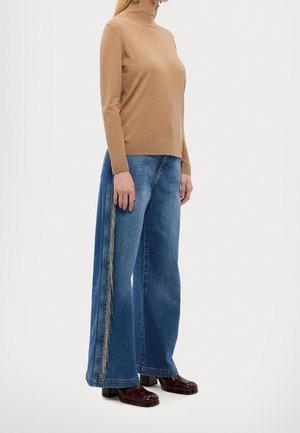 Beige Strick-Turtleneck-Pullover kombiniert mit weit geschnittenen blauen Denim-Jeans, die Fransenakzente an der Seite aufweisen. Dunkelbraune Absatzschuhe.