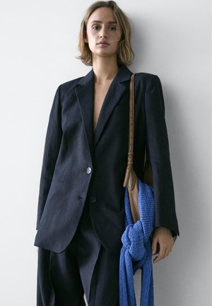 Kvinde med kort blondt hår iført oversized sort jakkesæt og bærer brun skuldertaske med blåt strikket stof på hvid baggrund.