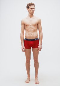 Νεαρός άνδρας με σγουρά μαλλιά, φορώντας κόκκινα μποξεράκια Polo Ralph Lauren, όρθιος χωρίς παπούτσια μπροστά σε απλό λευκό φόντο.