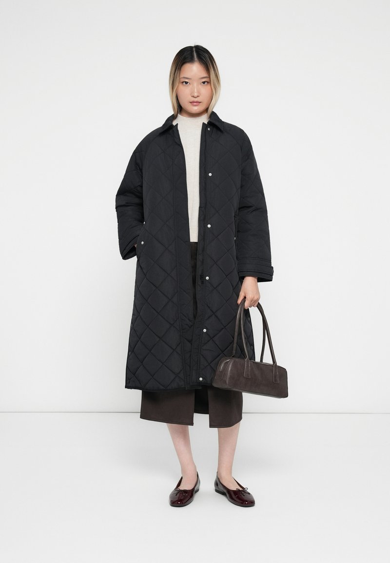 Zwarte gequiltte jas met drukknoopjes, lange mouwen en een rechte snit. Gepaard met een beige top, bruine rok en een kleine donkere tas.