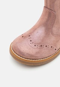 Różowe złote skórzane botki za kostkę z detalami w stylu brogue, okrągłym noskiem, jasnobrązową podeszwą oraz białymi akcentami wykończeniowymi. Gładka, metaliczna tekstura.