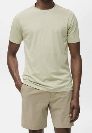 Mężczyzna ubrany w jasnobeżowy t-shirt z krótkim rękawem i okrągłym dekoltem oraz dopasowane beżowe szorty, stojący z rękami w kieszeniach na jednolitym tle.