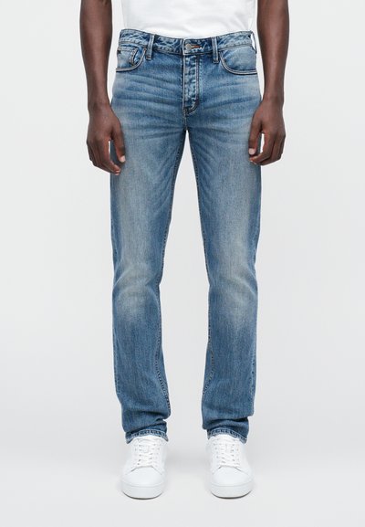 Šviesiai mėlyni džinsai, išskirtinio siauro kirpimo, su klasikinėmis penkiomis kišenėmis ir subtiliu nusidevėjimu, derinami su baltais sportiniais batais.