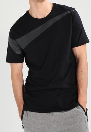 Mann trägt ein schwarzes Nike-T-Shirt mit großem dunkelgrauem Swoosh über der Brust und graue Jogginghose, Hände in den Taschen.