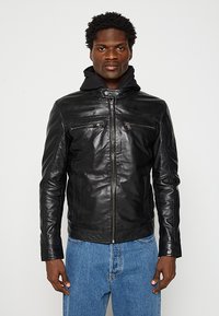 Schwarze Lederjacke mit Reißverschluss, zwei Brusttaschen und Kapuze. Glatte Textur und taillierte Passform, kombiniert mit blauen Jeans.