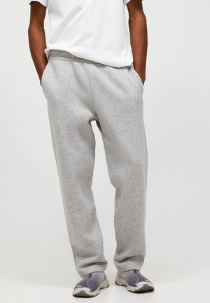 Person wearing light gray sweatpants, white shirt, and gray slip-on sneakers, standing with hands in pockets against a plain background.