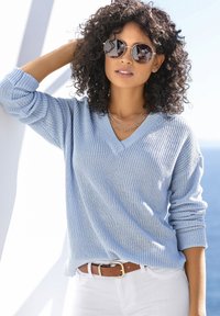 Hellblauer Strickpullover mit V-Ausschnitt, versehen mit rippstruktur und langen Ärmeln, kombiniert mit einem braunen Gürtel und weißen Hosen.