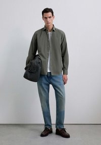Camisa verde de botón sobre una camiseta blanca, combinada con jeans azul claro y zapatos marrón oscuro. Sosteniendo una bolsa gris oscura. Fondo minimalista.