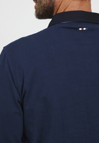 Námornícky polo tričko s tmavým golierom. Na zadnej strane má malý trojfarebný horizontálny pruh. Hladká bavlnená tkanina.