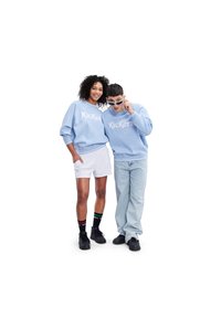 Due modelli indossano felpe azzurre chiare con il logo "Kickers", abbinate a pantaloncini bianchi e jeans. Sneakers nere e calzini a fantasia completano il look.