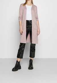 Lång, ljusrosa cardigan med öppen framsida, två fickor, bärs över en vit topp och blanka svarta cropped byxor. Svarta kraftiga ankelstövlar fulländar looken.