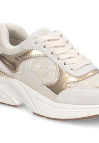 Zapatilla blanca y dorada con suela gruesa, paneles de malla y ante, cordones beige y un logo decorativo en el lateral.