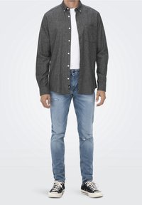 Chemise grise à boutons en tissu texturé, portée sur un t-shirt blanc, associée à un jean en denim bleu clair déchiré et des baskets noires.