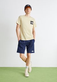 T-shirt in cotone beige con una patch nera, abbinata a pantaloni corti navy e sneaker crema. Il modello indossa un berretto scuro. Sfondo verde.