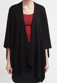 Vrouw in een losse, open voorkant zwarte cardigan over een rode tanktop en zwarte rok, staande tegen een eenvoudige witte achtergrond.