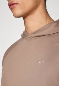 Καφέ φούτερ με κουκούλα από απαλό ύφασμα, που διαθέτει ένα μικρό, κεντητό λογότυπο της Nike σε λίγο πιο σκούρο τόνο. Η γραμμή είναι εφαρμοστή.