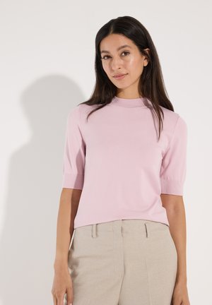 Junge Frau mit langen dunklen Haaren, die ein hellrosa Kurzarmtop und beige Hosen vor einem schlichten hellen Hintergrund trägt.