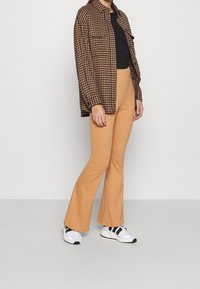 Bruine houndstooth oversized shirt, zwarte top, geribbelde crèmekleurige flared broek en grijze sneakers met zwarte accenten. Simpele, moderne outfit.