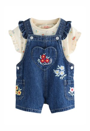 Kind draagt een spijkerjurk met bloemrijke borduursels, gele sokken en een bloemetjesblouse, staand bij beschilderde bloemversieringen.