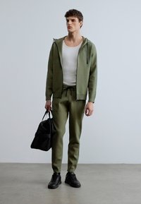 Sudadera con capucha y pantalones deportivos de punto en verde oliva, con detalles acanalados, combinados con una bolsa negra y zapatillas negras, sobre un fondo neutro.