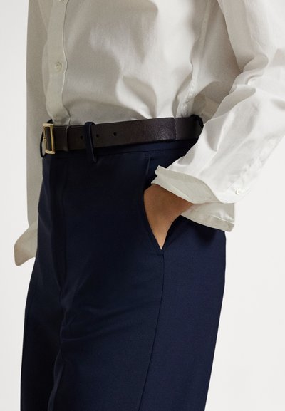 Camisa branca com botões e mangas arregaçadas, combinada com calças azuis-escuro e um cinto de couro castanho escuro com fivela dourada.