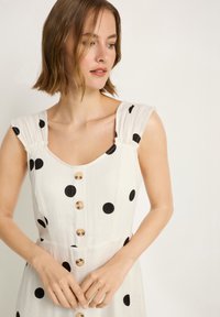 Weißes Kleid mit schwarzen Tupfen, quadratischem Ausschnitt und Knopfleiste vorne. Mit plissierten Trägern und leichtem Stoff.