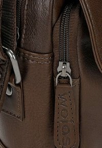 Dettaglio di una borsa in pelle marrone con una tasca zip, accessori in argento e logo in rilievo su un'etichetta in pelle. Texture liscia con cuciture visibili.