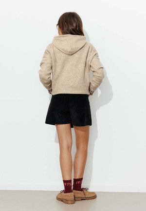 Femme vue de dos portant un sweat à capuche beige, un short noir, des chaussettes bordeaux avec « 1975 », et des chaussures à lacets couleur beige sur fond blanc.