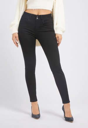 Femme portant un jean skinny noir taille haute, un pull court en laine blanc pelucheux et des chaussures à talons hauts scintillantes de couleur sombre, debout sur fond blanc.