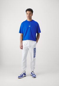 Blaues Baumwoll-T-Shirt, weiße Jogginghose mit blauem "HUGO"-Logo und blau-weißen Turnschuhen. Das Model steht vor einem neutralen Hintergrund.
