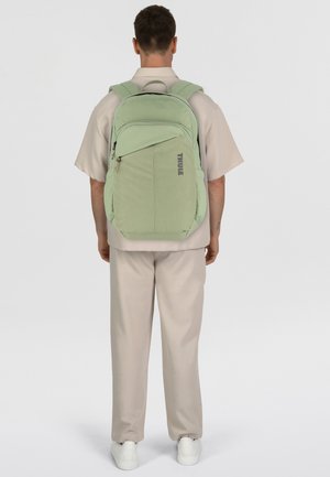 Sac à dos vert clair avec des bretelles rembourrées, plusieurs poches zippées et un logo "THULE" gris proéminent à l'avant.