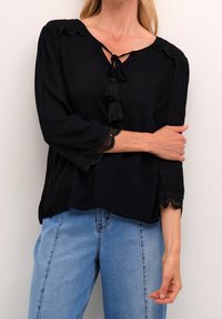 Svart blus med trekvartslånga ärmar, förskönad med spetsdetaljer och en knytning med tofsar vid kragen. Matchad med ljusblå jeans.