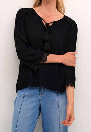 Schwarze Bluse mit Dreiviertelärmeln, ausgestattet mit Spitzendetails und einer Bindeschleife mit Quasten am Hals. Kombiniert mit hellblauen Jeans.