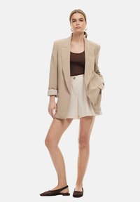Blazer beige surdimensionné avec des manches retroussées, débardeur marron à rayures, short plissé crème et chaussures à enfiler en tissage foncé.