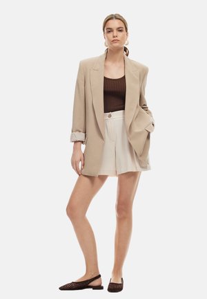 Beige oversized blazer met opgerolde mouwen, bruine geribbelde tanktop, crèmekleurige geplooide shorts en donkergeweven instapschoenen.