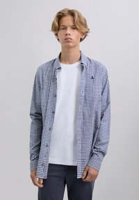 Joven con cabello rubio que lleva una camisa de cuadros azul con botones sobre una camiseta blanca y pantalones oscuros, contra un fondo claro y liso.