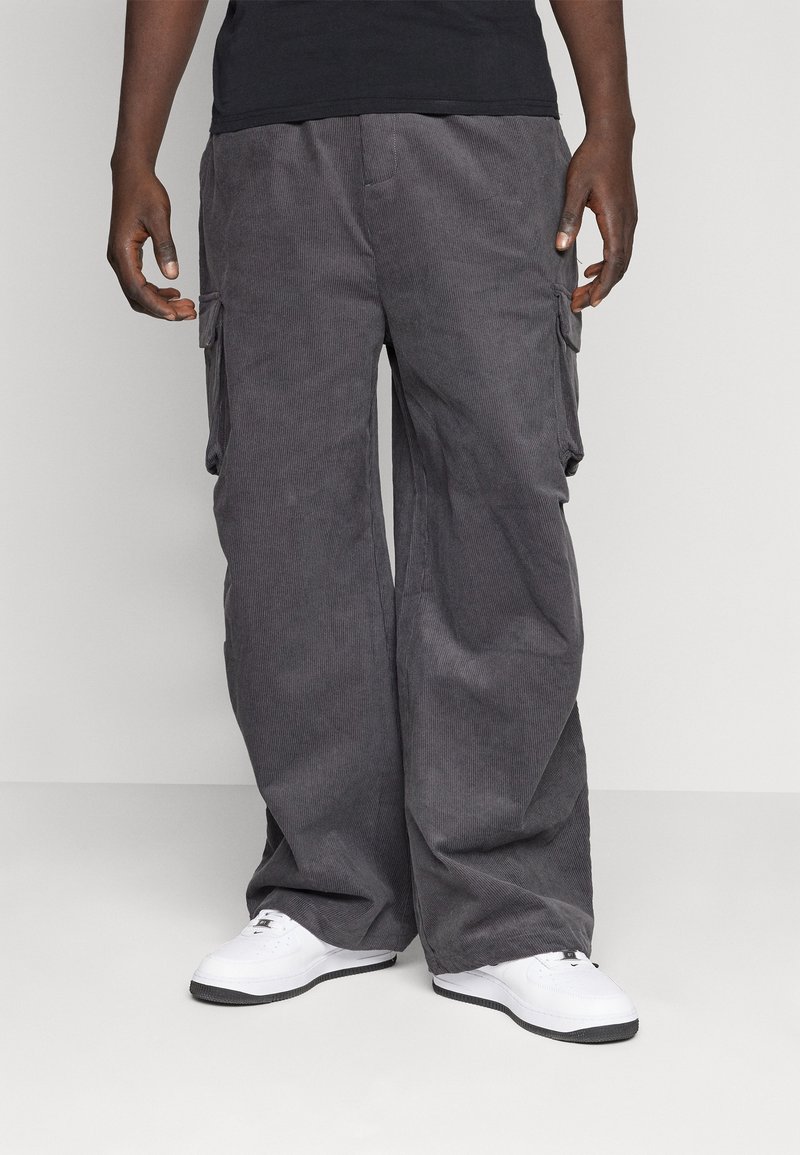 Homme portant un pantalon cargo gris à jambes amples et des baskets blanches, debout devant un fond uni.