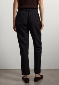 Persona che indossa pantaloni neri su misura fino alla caviglia e scarpe flat marroni, in piedi contro un muro chiaro e semplice.