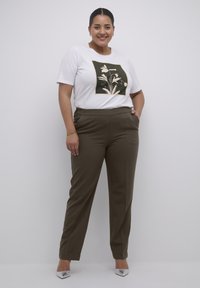 Weißes Baumwoll-T-Shirt mit grafischem Druck in dunklem Grün und Blumenmuster, kombiniert mit braunen, maßgeschneiderten Hosen und silbernen, spitzen Schuhen.