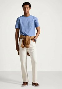 Νεαρός άνδρας στέκεται φορώντας μπλε μπλούζα, λευκά παντελόνια, καφέ πουλόβερ δεμένο γύρω από τη μέση, καφέ αθλητικά παπούτσια και ρολόι στον αριστερό καρπό.