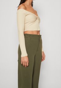 Beige žebrovaný top s odhalenými rameny a dlouhými rukávy, s twistem v přední části, kombinovaný s olivově zelenými kalhotami s vysokým pasem, které mají zip a boční kapsy.