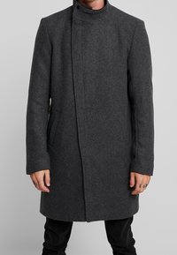 Cappotto di lana grigio con colletto alto, design slanciato, chiusura frontale nascosta e tasche laterali. Maniche lunghe con taglio dritto.
