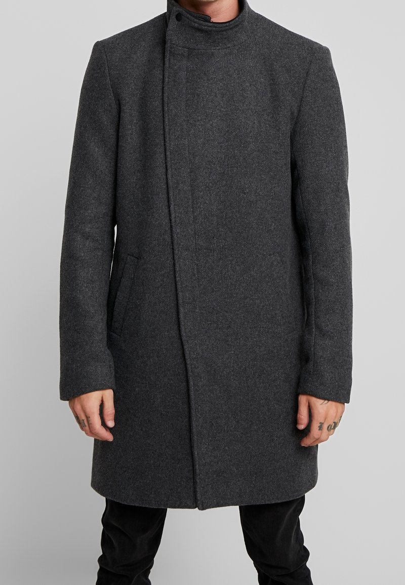 Cappotto di lana grigio con colletto alto, design slanciato, chiusura frontale nascosta e tasche laterali. Maniche lunghe con taglio dritto.