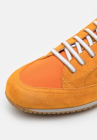 Gros plan d'une basket orange avec une texture en daim, des lacets blancs et une semelle en caoutchouc sur fond blanc.