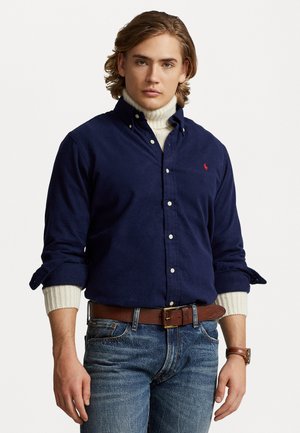 Camicia button-down in corduroy blu navy con bottoni bianchi, caratterizzata da maniche arrotolate su un dolcevita crema, abbinata a una cintura in pelle marrone e jeans di denim.